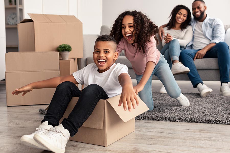 Personal Insurance - Kids Pushing Each Other Around a Room in Cardboard Moving Boxes, Their Parents Smiling on the Couch Behind Them